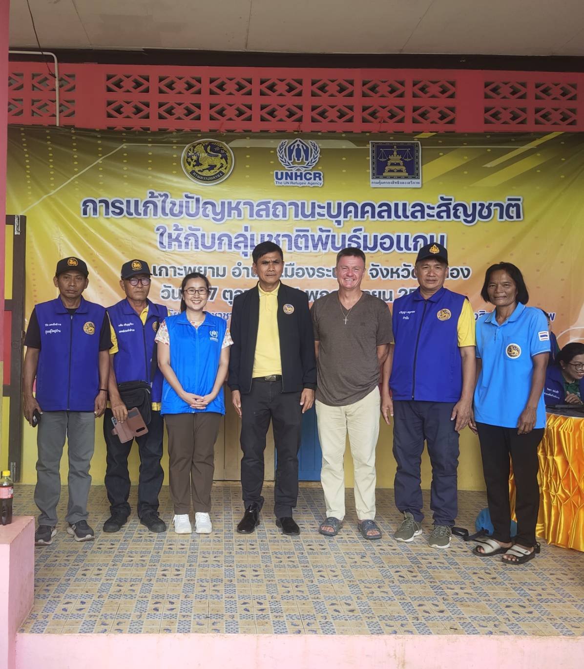 A group of adults, among them officials in blue vests, a UNHCR staff member and a Western man, in front of a UNHCR and Thai government banner at a Moken ID registration drive on Koh Phayam in 2024