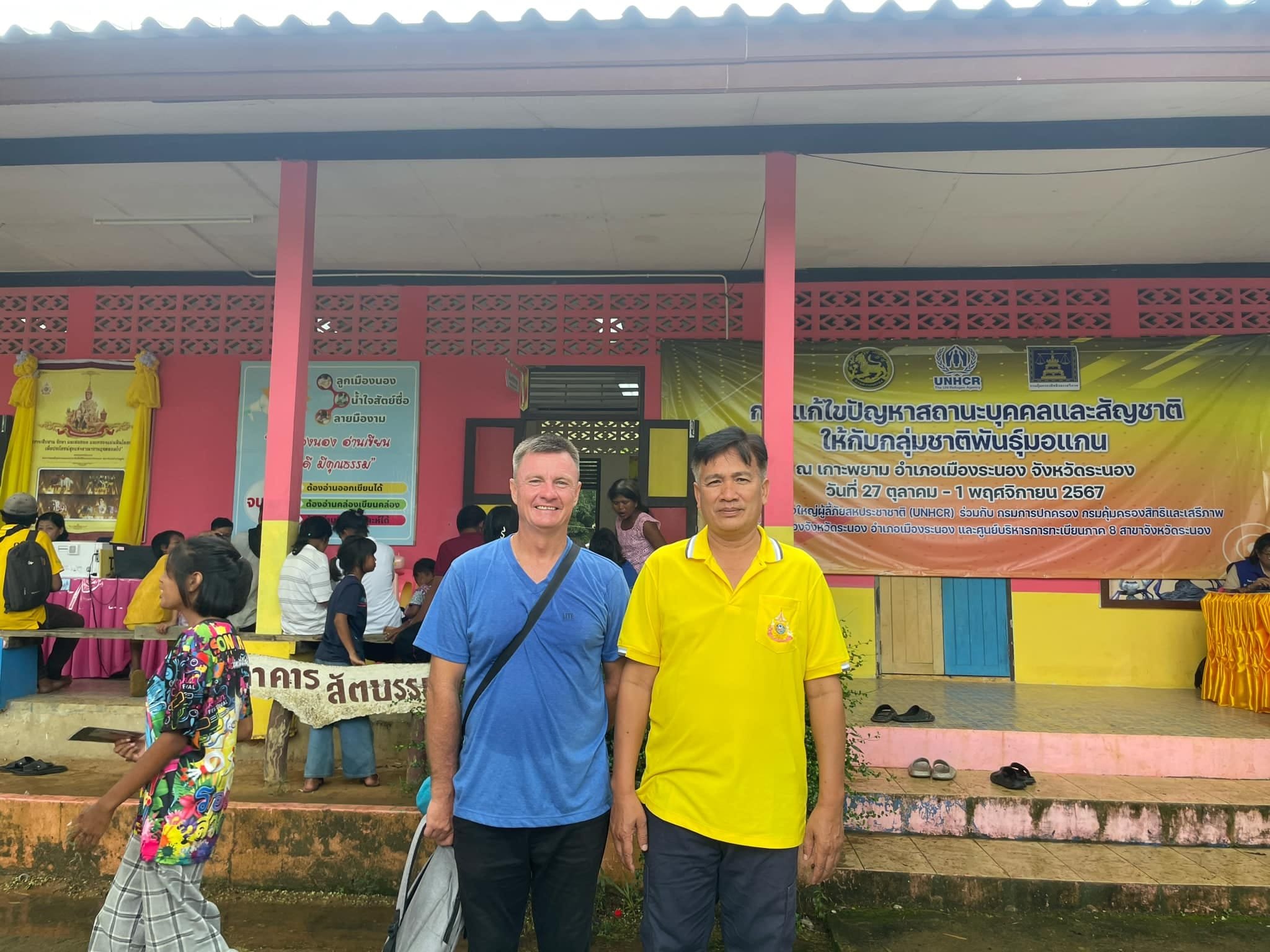 Philip with the island headman in a yellow shirt in front of a school building on Koh Phayam, under a UNHCR and Thai government banner for the 2024 Moken registration drive, with people waiting at tables behind them