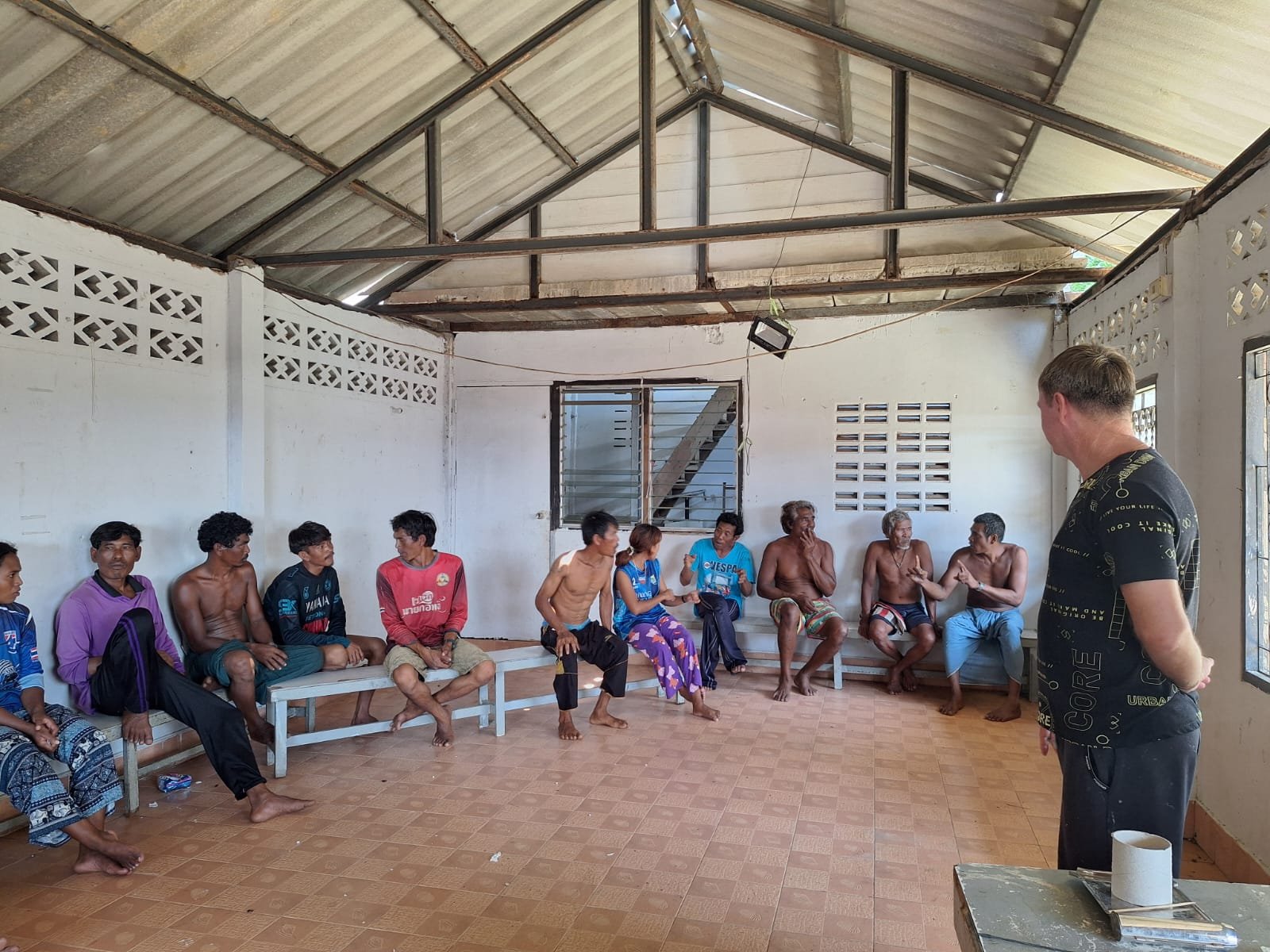 About a dozen Moken adults seated on benches around a bare community hall on Koh Chang, one gesturing as he speaks, while a man stands with his back to the camera at the July 2026 schooling meeting
