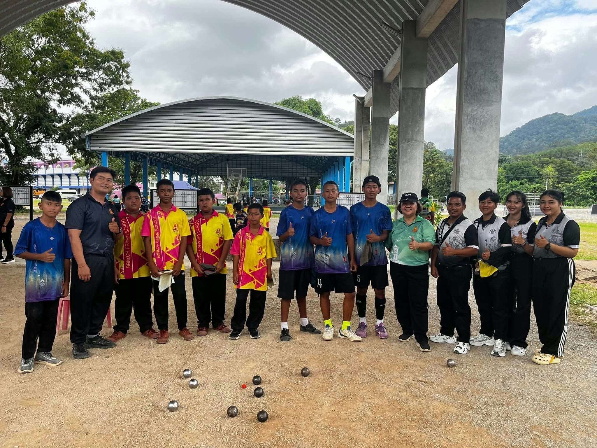 Two school petanque teams with their coaches and officials beside a petanque pitch under a large covered sports arena; the Koh Phayam school boys, in pink and yellow, came second of 16 schools