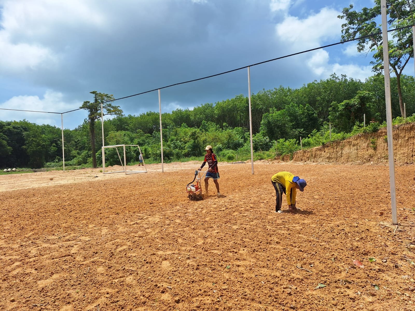 Two young men planting grass on a quarter of the bare red-earth soccer field in July 2025, one working the soil with a small tiller while the other plants by hand, with the tall net poles, a goal and forest behind