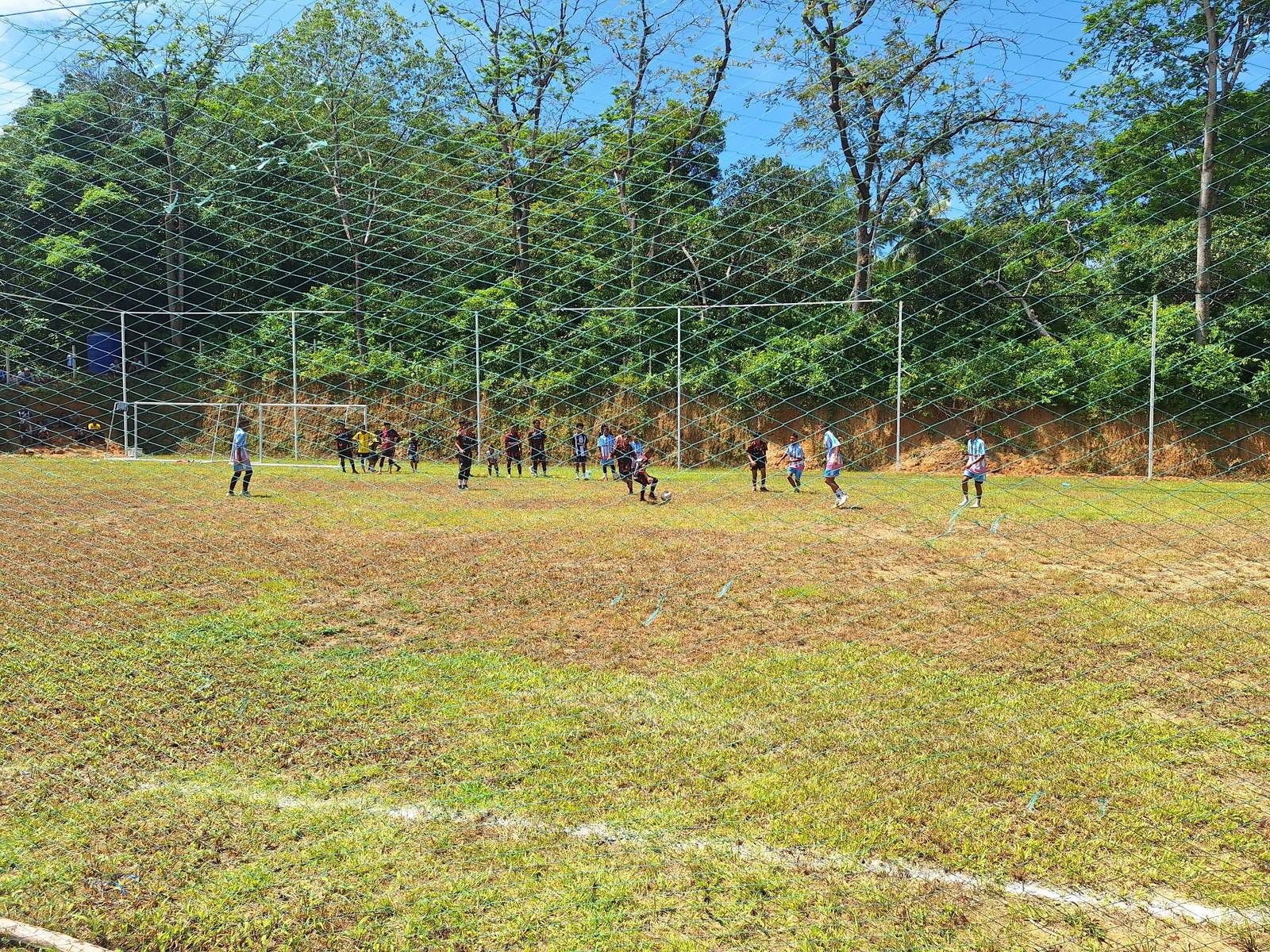 Young players in two kits contesting the ball in midfield on the now-grassed soccer field, seen through the perimeter net, on the day of the Phayam Cup