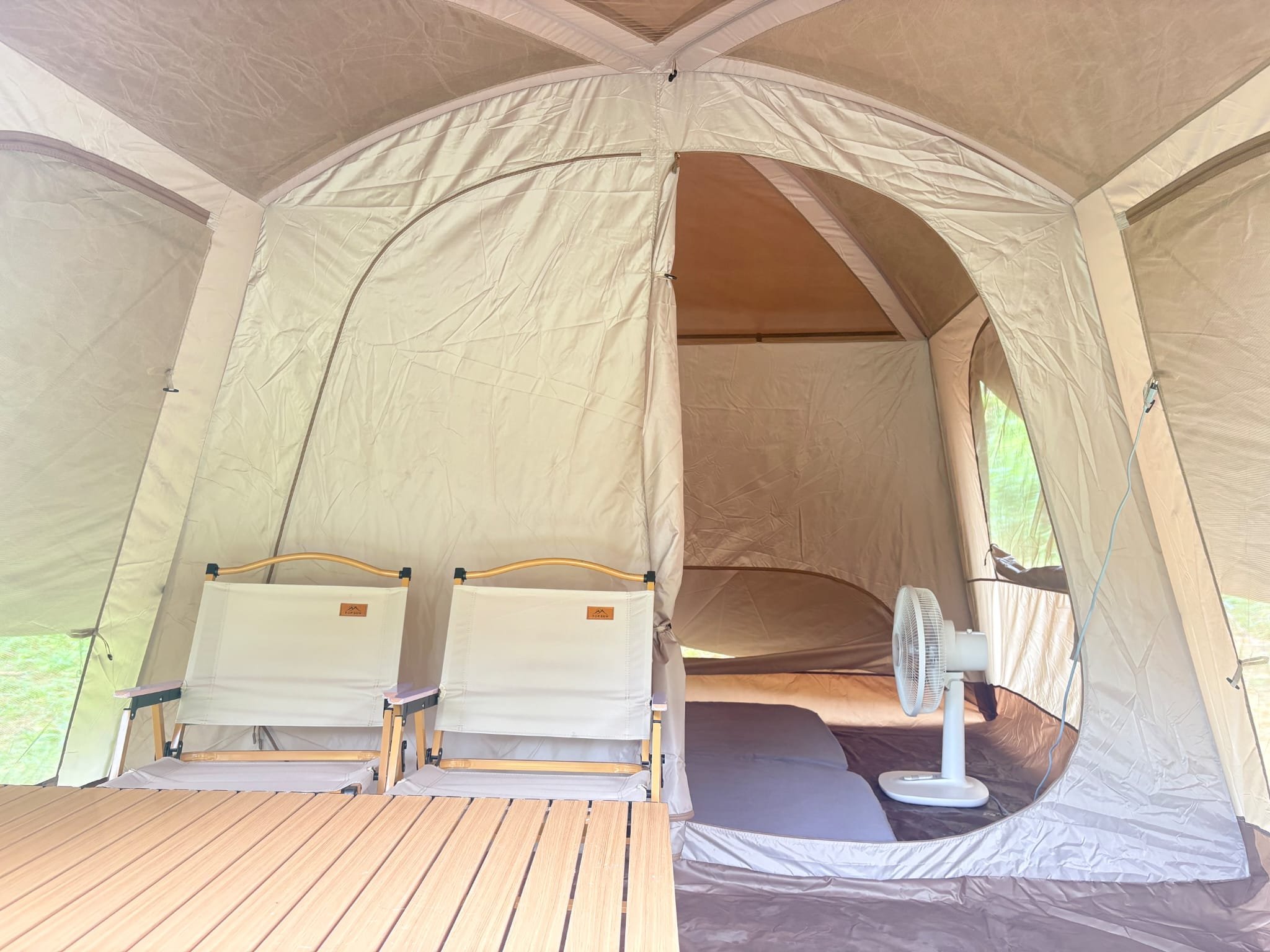 Inside the tent: two folding canvas chairs and a slatted wooden table in the living area, with the sleeping compartment, mattresses and a fan behind