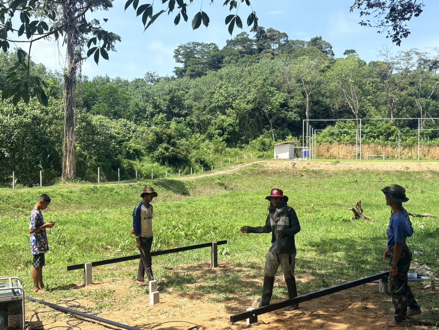 Four people starting a tent platform on cleared ground: black steel beams resting on small concrete footings, with the fenced soccer field on the slope behind