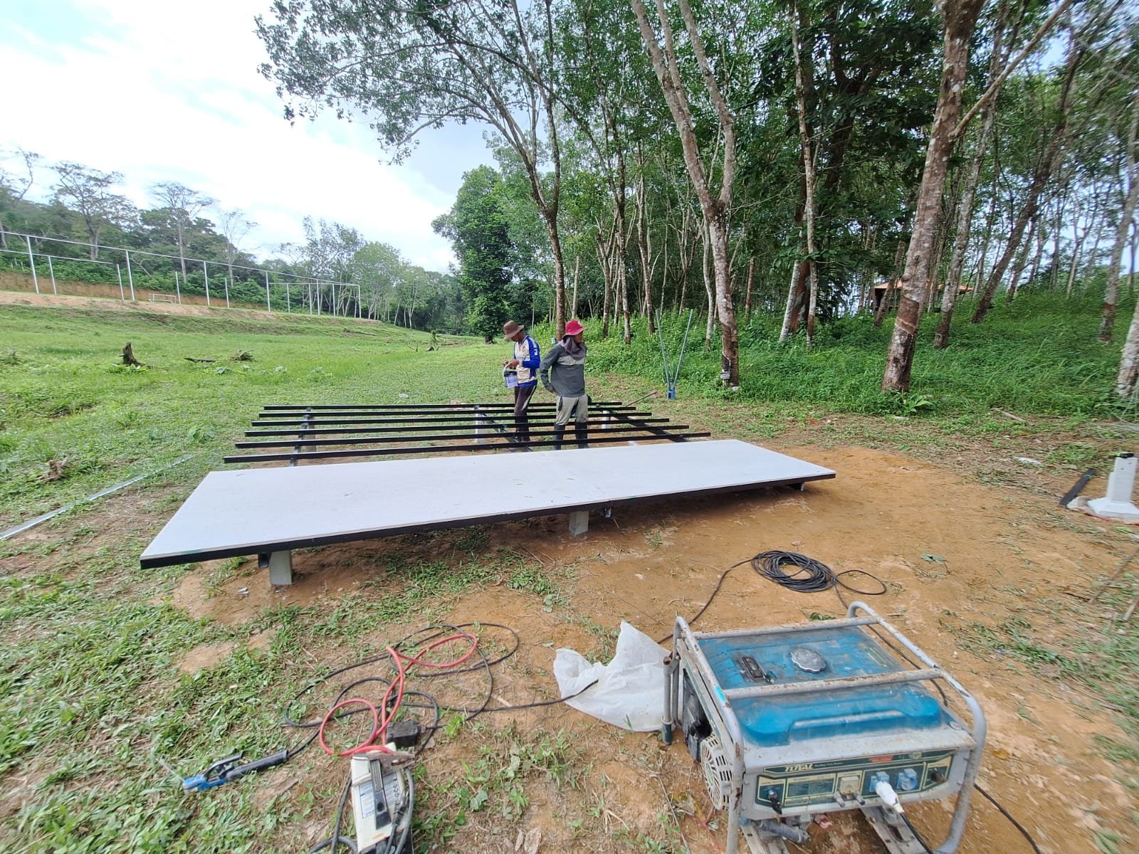 Two workers standing among the bare steel beams of a platform frame, beside a platform already finished with a smooth pale deck on concrete posts; a generator and welding cables lie in the foreground