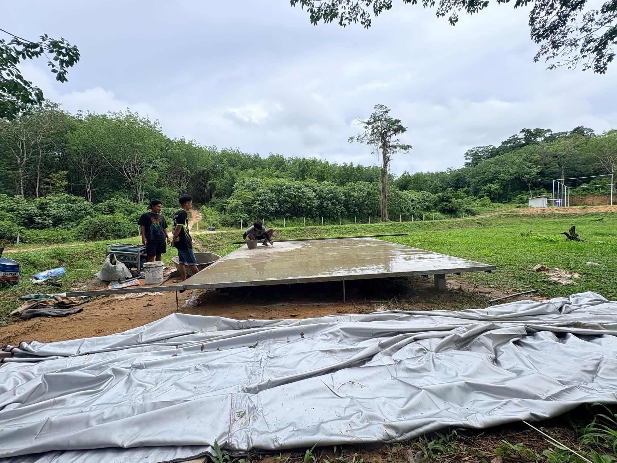 Wud and the others poured the rest of the cement: a young man crouches by a bucket smoothing the wet surface of the raised platform while two others look on, the tent canvas spread out on the grass in front
