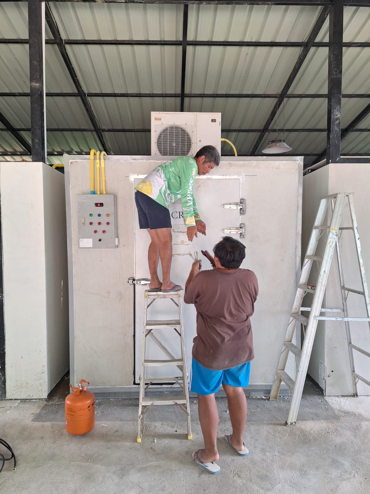 Two men fitting the door of the newly installed walk-in cold room for ice, one standing on a stepladder, with the cooling unit on top