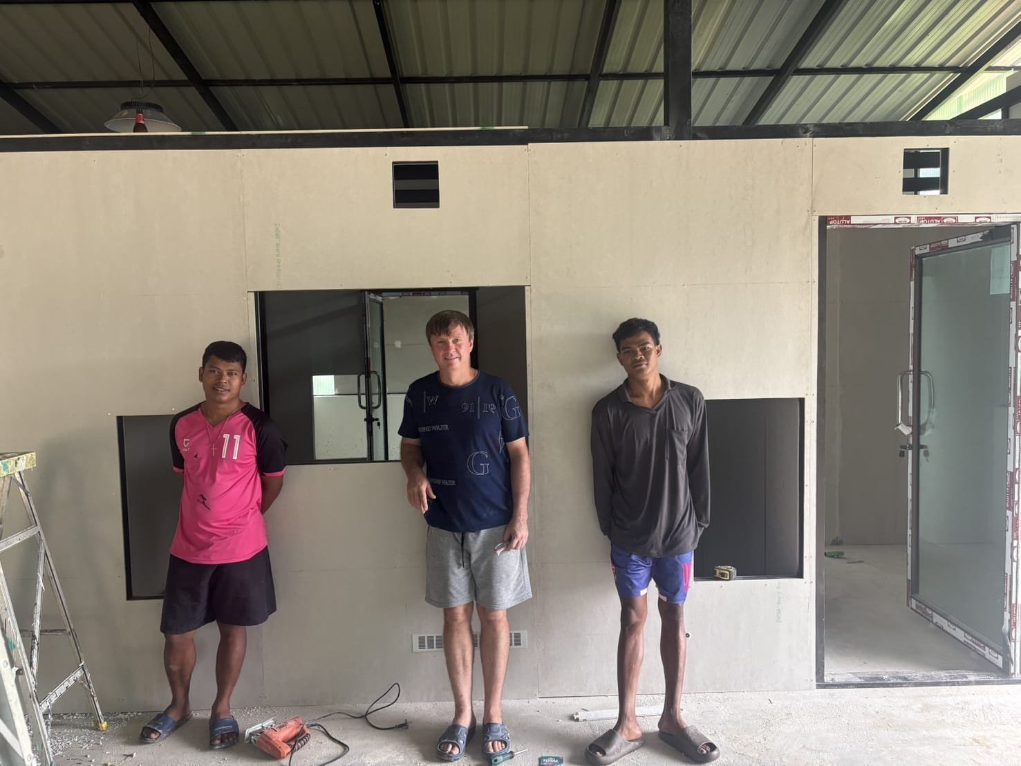 Three men standing in front of new board partition walls with window openings and a glass door, as the water-bottling rooms are built inside the factory shed