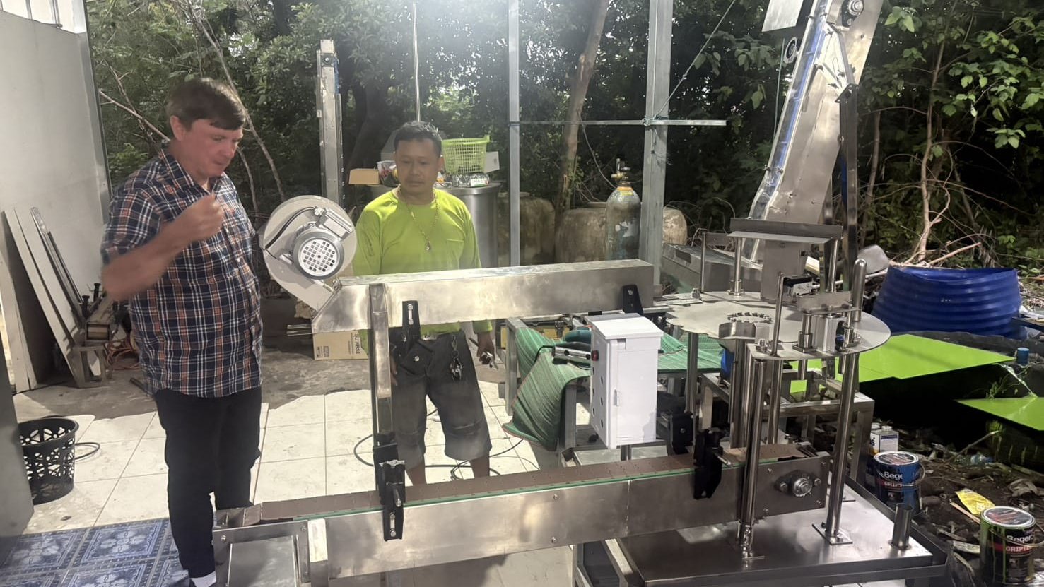 Philip inspecting a stainless-steel bottle-capping machine with another man at an outdoor workshop, on a November 2025 trip to look at water machines for the factory