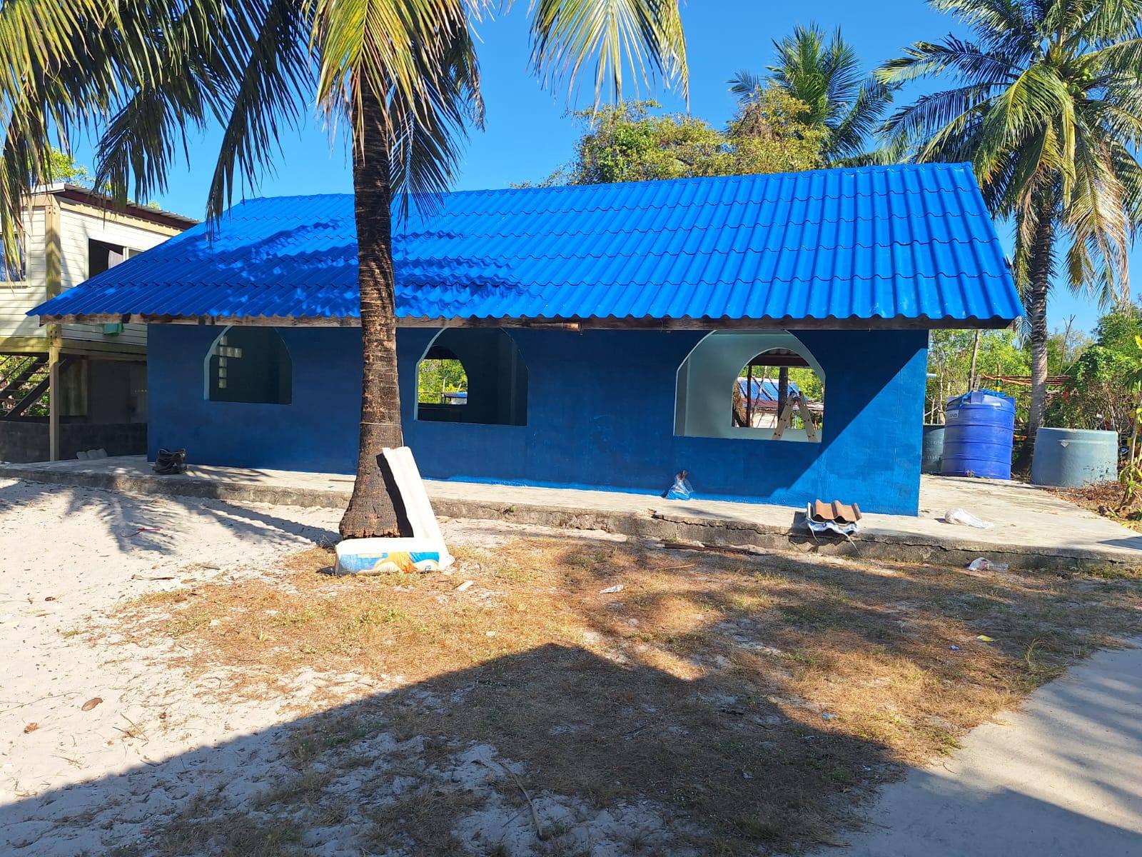 The small church freshly painted bright blue under a new blue corrugated roof, with arched window openings (a white tile cross visible on the inside wall through one of them), among palm trees and water tanks