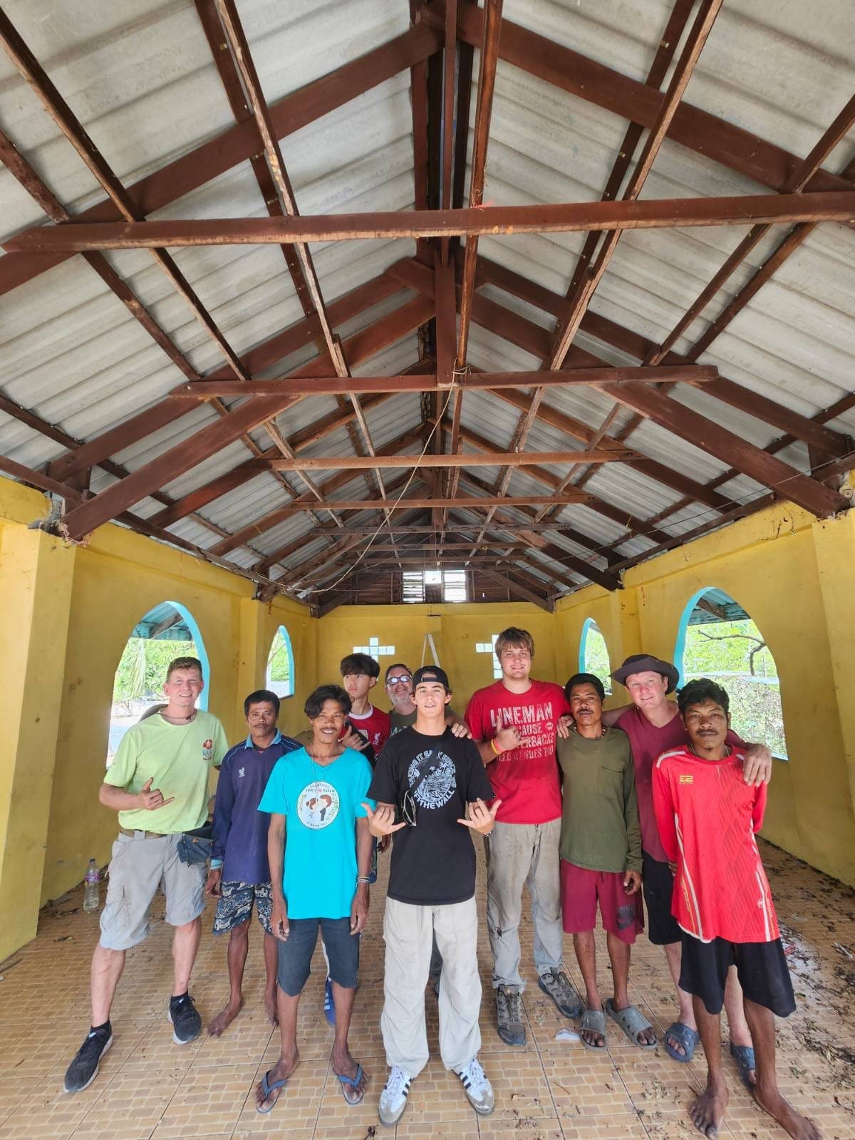 About ten men, volunteers from Kauai and local men, standing together inside the yellow church under its exposed timber roof trusses during the January 2026 renovation