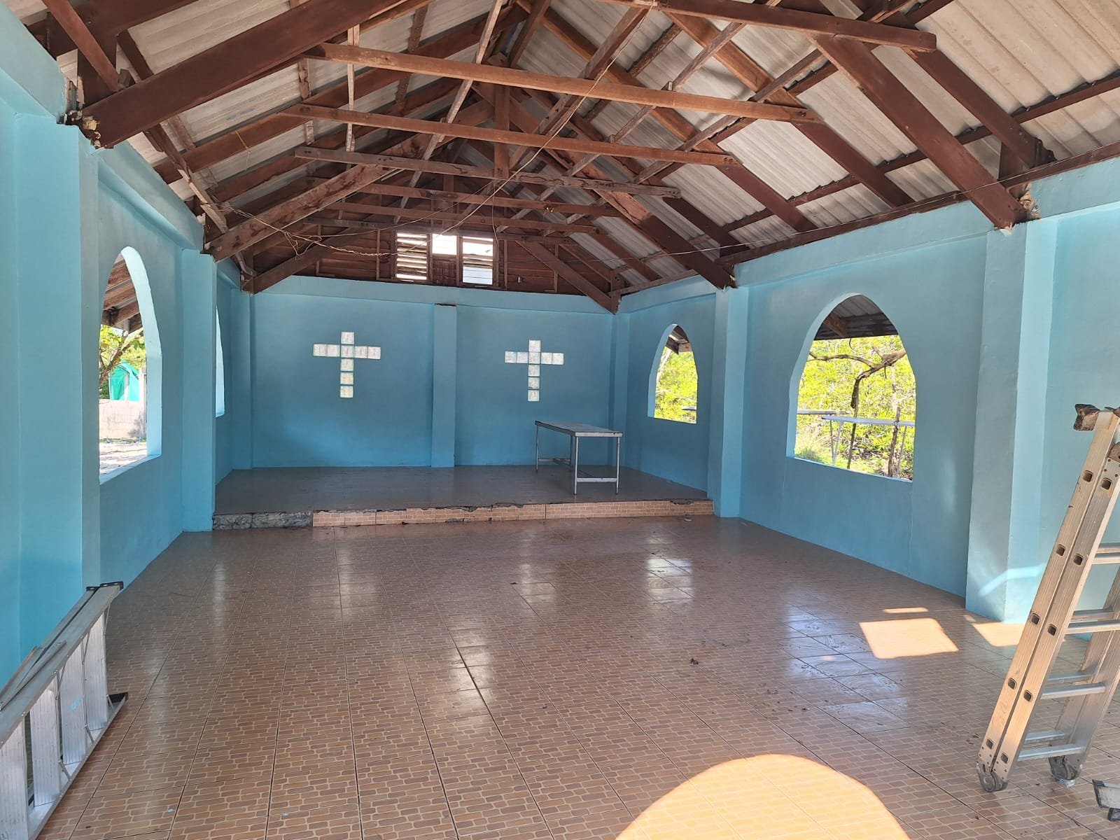The empty church interior after repainting: light-blue walls with two white tile crosses, arched windows, a tiled floor and timber roof trusses, with ladders still standing