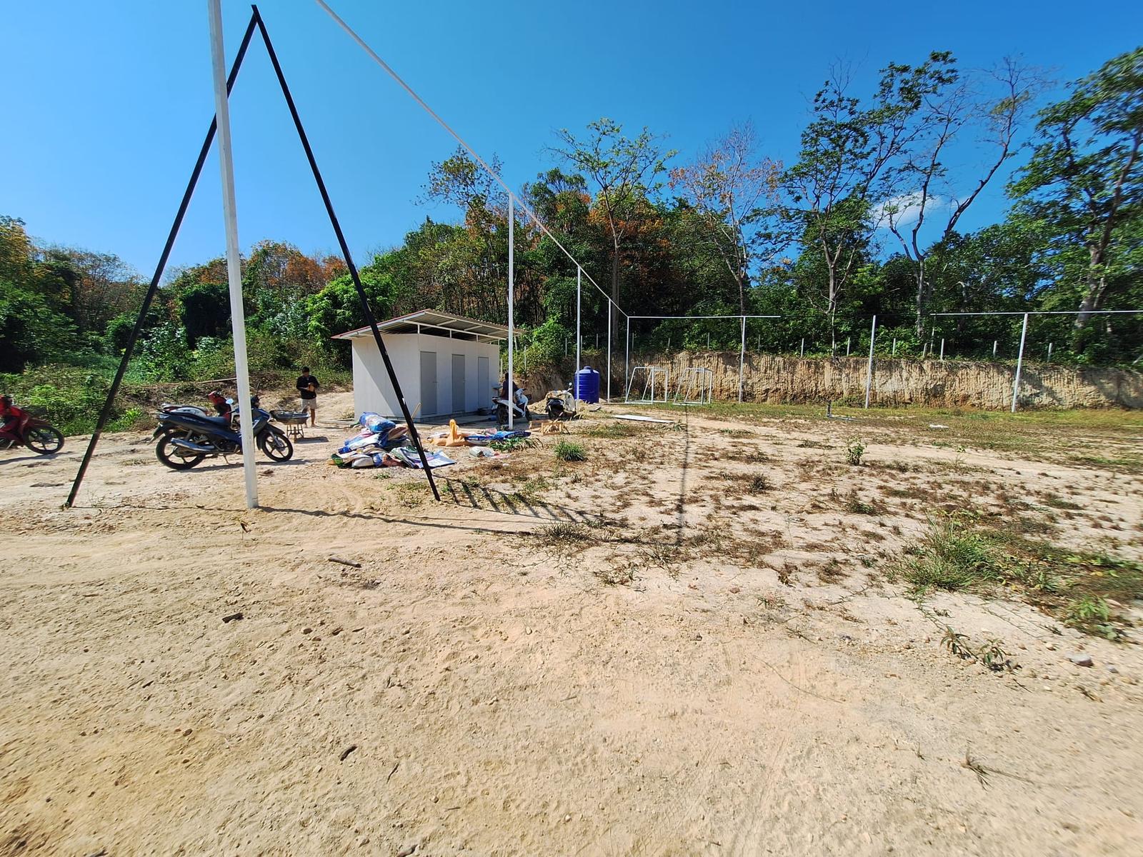 A small white bathroom block beside the sandy soccer field, with the tall white poles for the perimeter netting