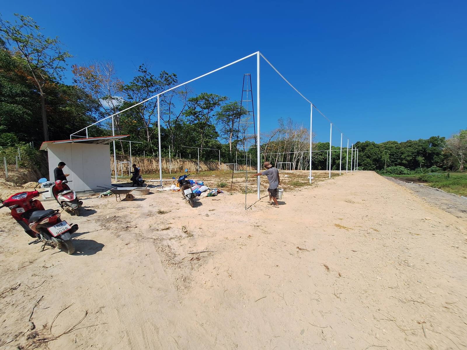 The sandy soccer field with its white netting poles, goal frames and new bathroom block, with two men working on site