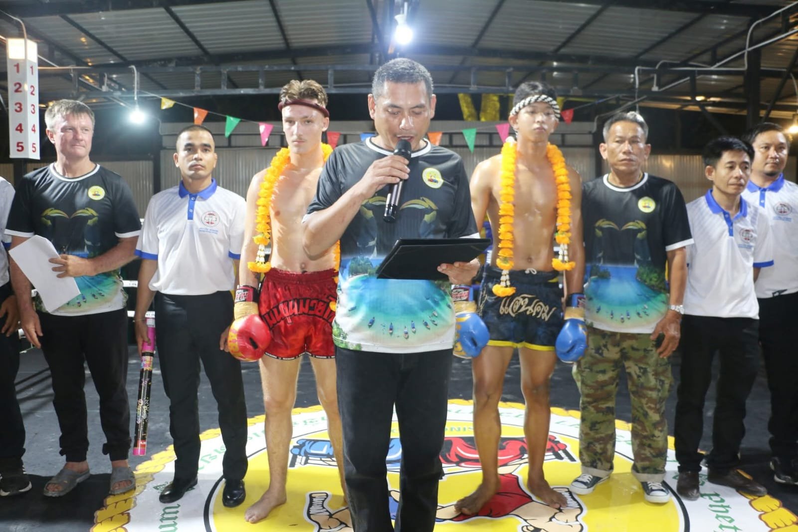 Officials in matching shirts in a Muay Thai ring with two garlanded fighters, as one man reads an announcement into a microphone