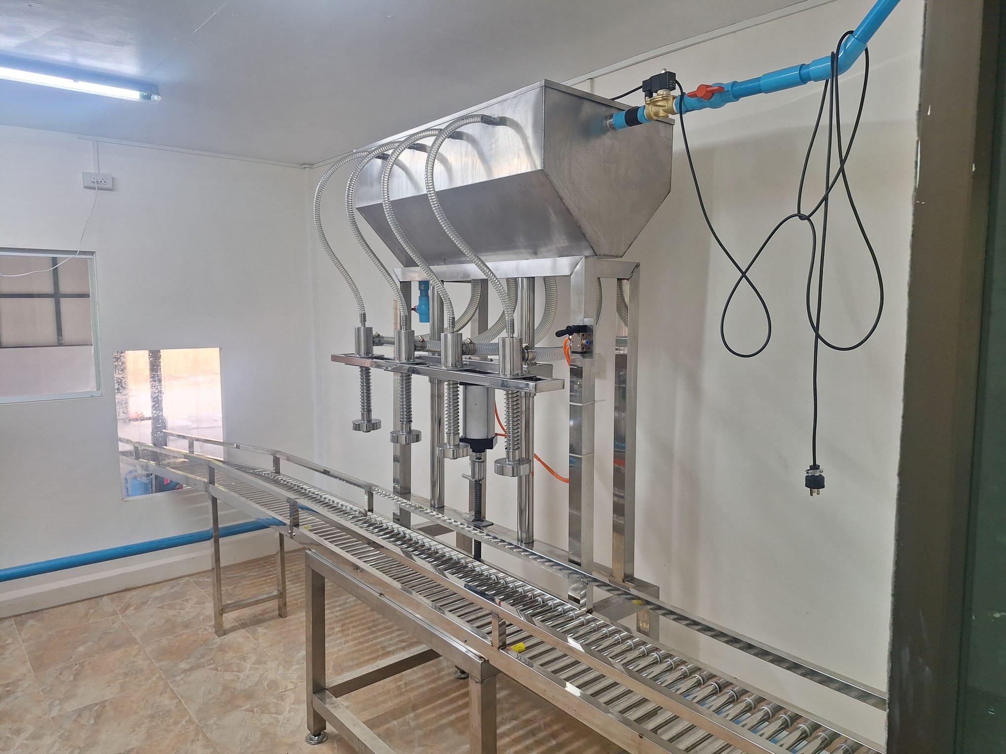 A four-head stainless-steel water-filling machine above a roller conveyor in the factory's white clean room