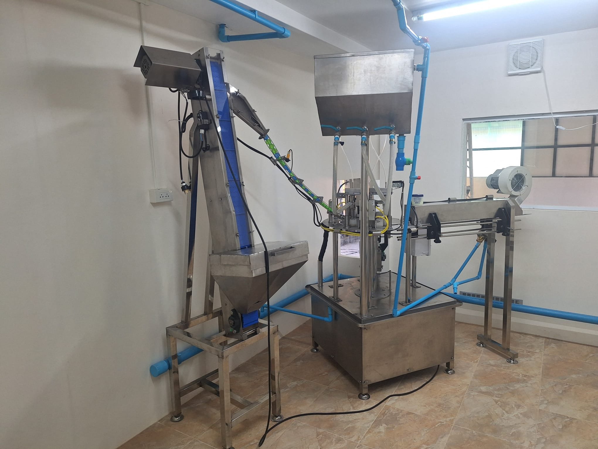 The rotary water-bottle filling and capping machine with its blue cap conveyor, plumbed in with blue pipes in a white tiled room
