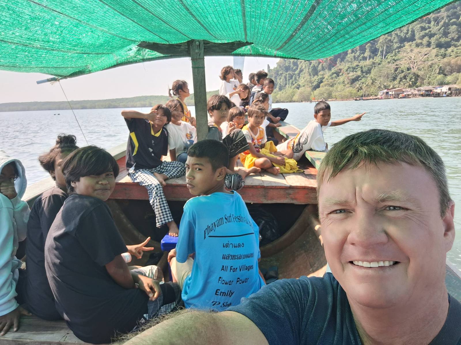 A selfie on a shaded long-tail boat packed with around twenty children, crossing toward a forested island with a stilt village