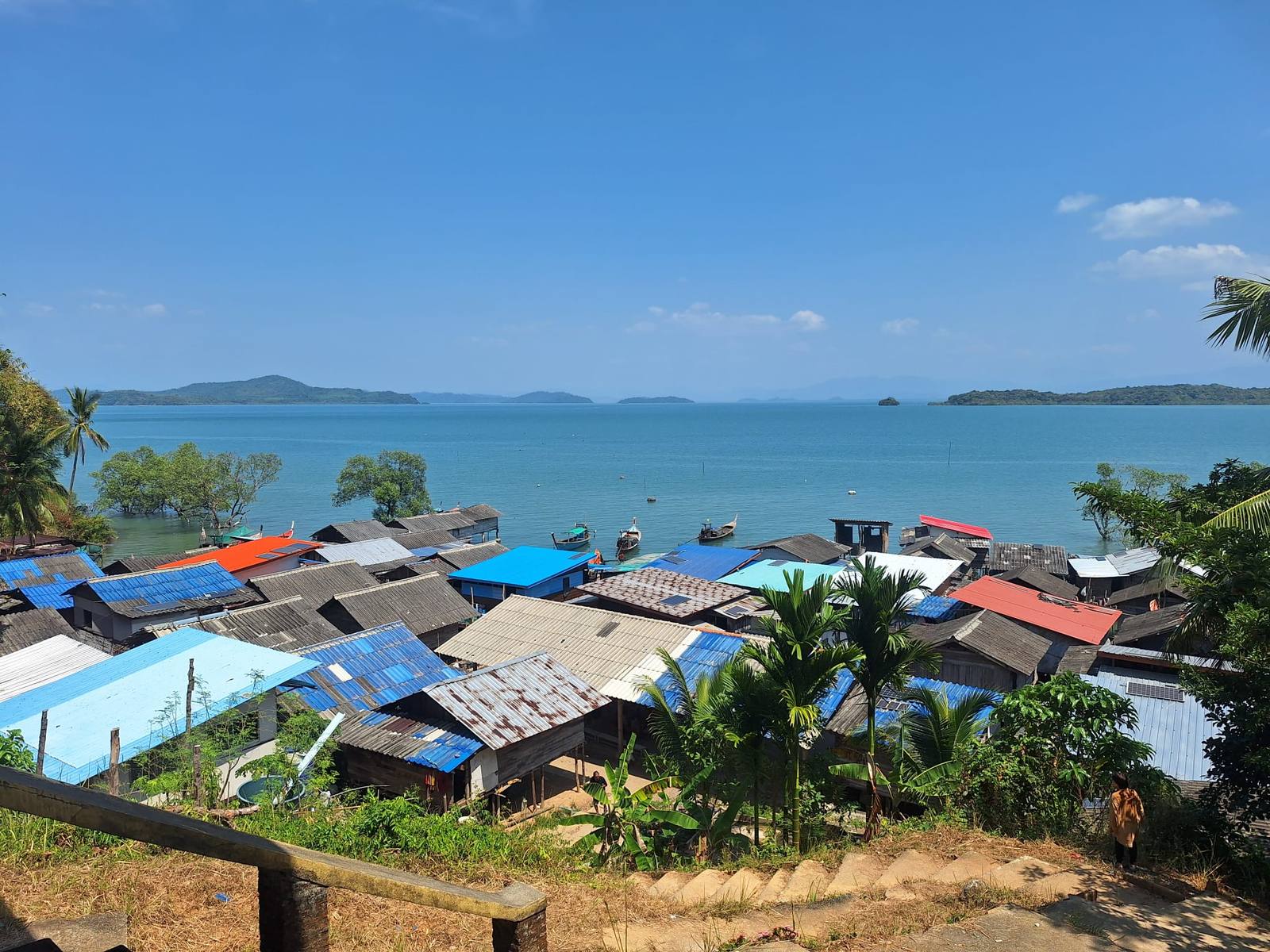 View over the colourful tin roofs of a coastal village, with long-tail boats moored in a calm bay and islands on the horizon