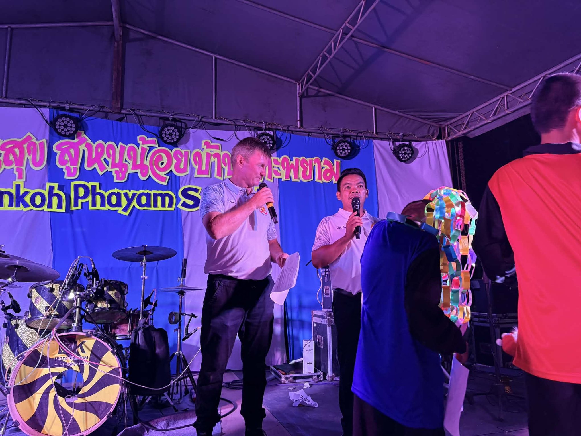 Two MCs with microphones on stage under the Koh Phayam school banner as costumed performers come onto the stage