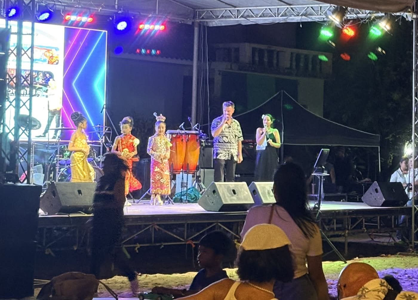 Two MCs with microphones on a lit outdoor festival stage at night, beside three young girls in traditional Thai costume