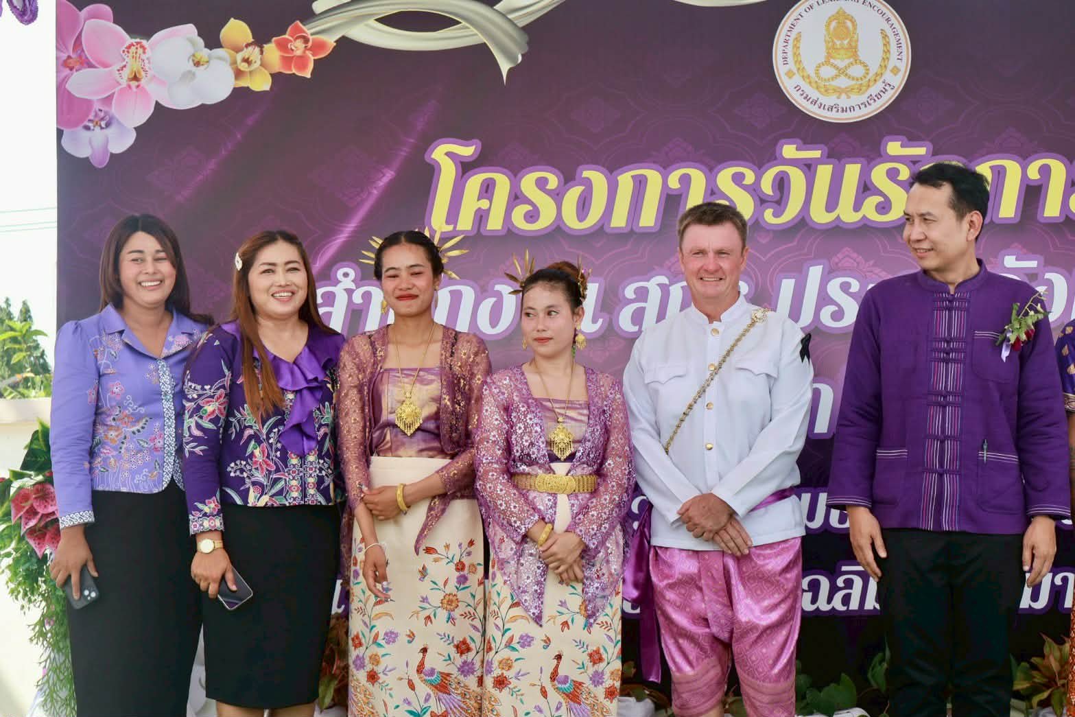 Six adults, two women in traditional Thai dress and a Western man in a white jacket and pink Thai trousers among them, in front of a purple event banner