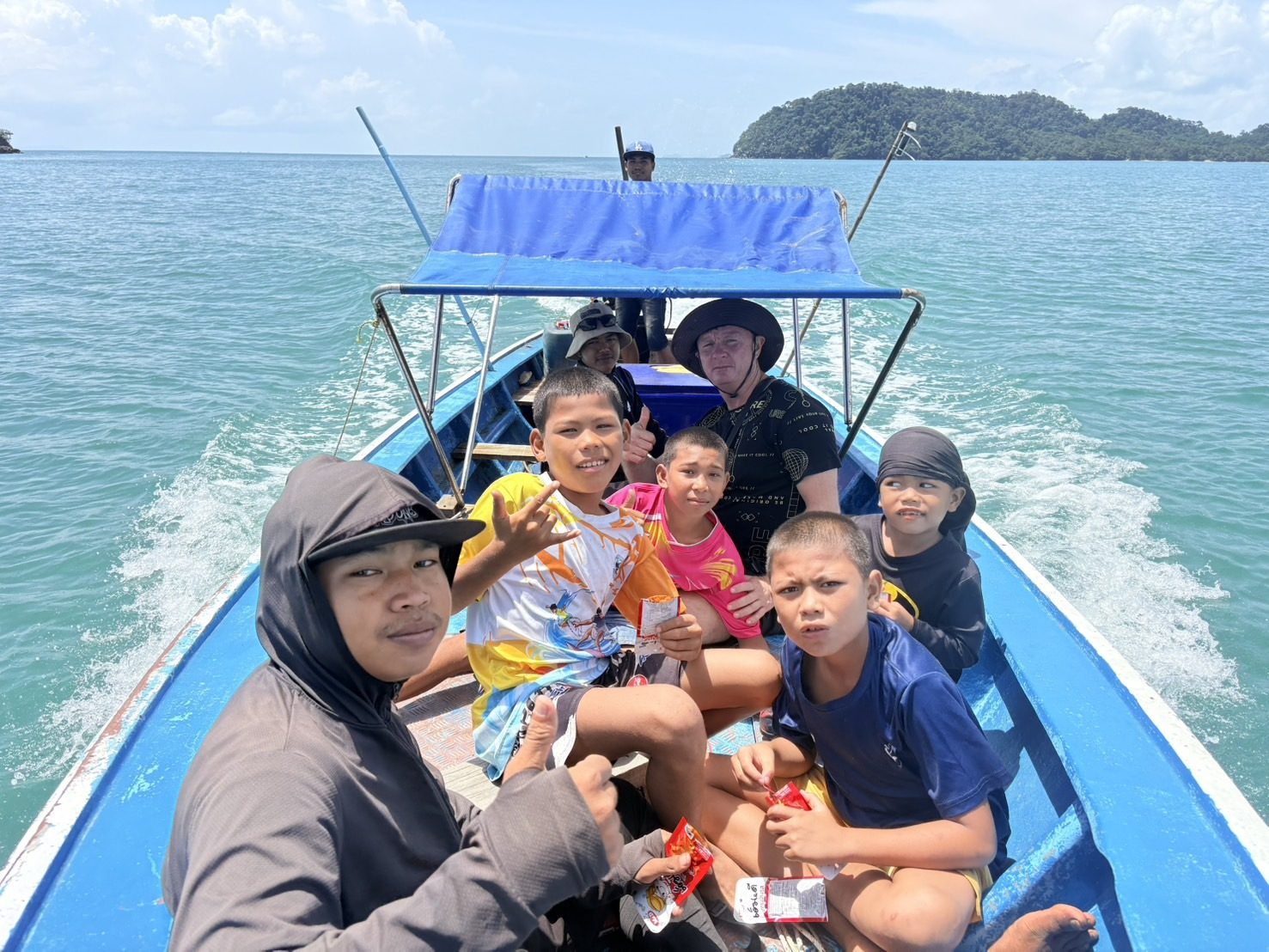 On the way to Koh Chang: a man in a bush hat with Moken boys and young men in a blue longtail boat, crossing turquoise sea toward a forested island