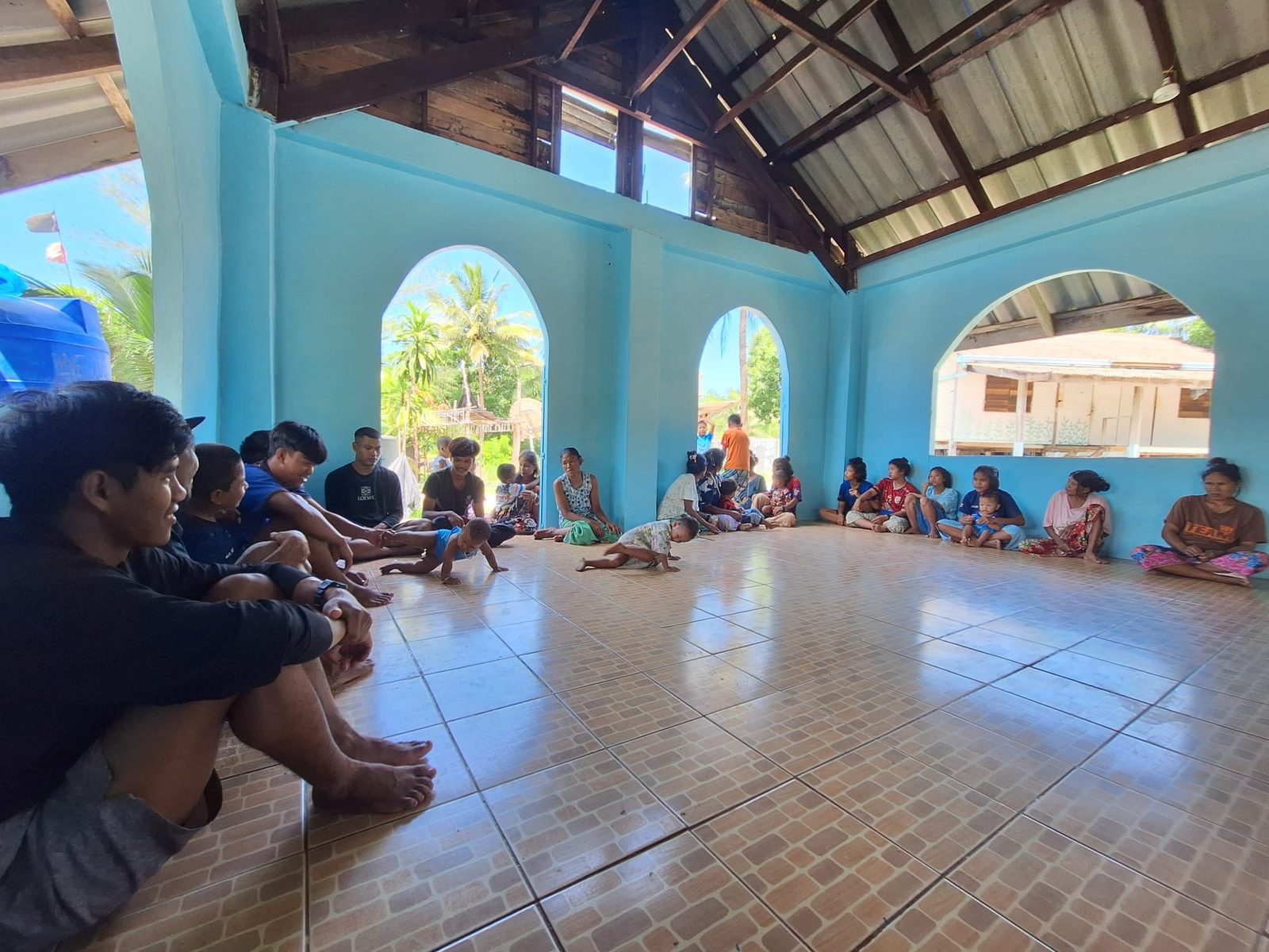 Moken young men, women and toddlers seated around the walls of the turquoise church hall, with arched windows open to the palms