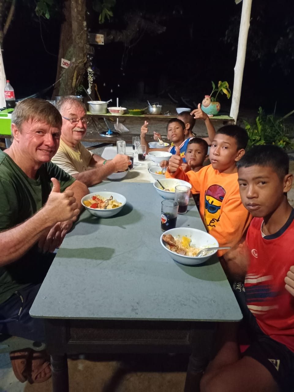 Two men and five Moken boys at an evening meal at an outdoor table during the August visit, several giving a thumbs-up