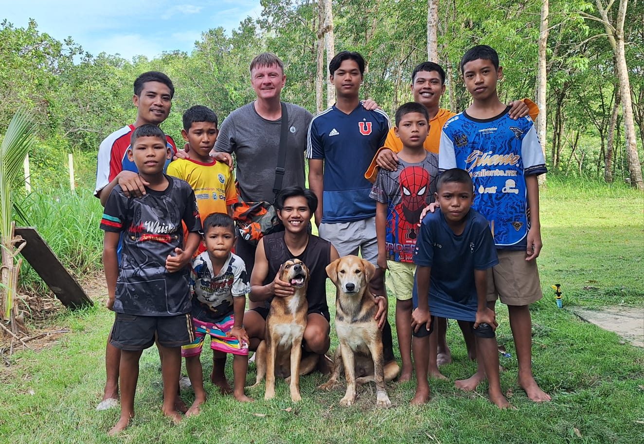 A man with about ten Moken boys and young men and two dogs on a grassy clearing with trees behind, all smiles