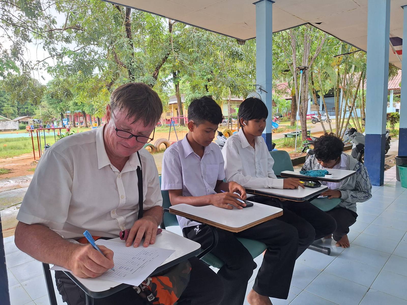 Exam day: Philip writing an exam at a tablet-arm desk on a school veranda, beside teenage students in school shirts doing the same