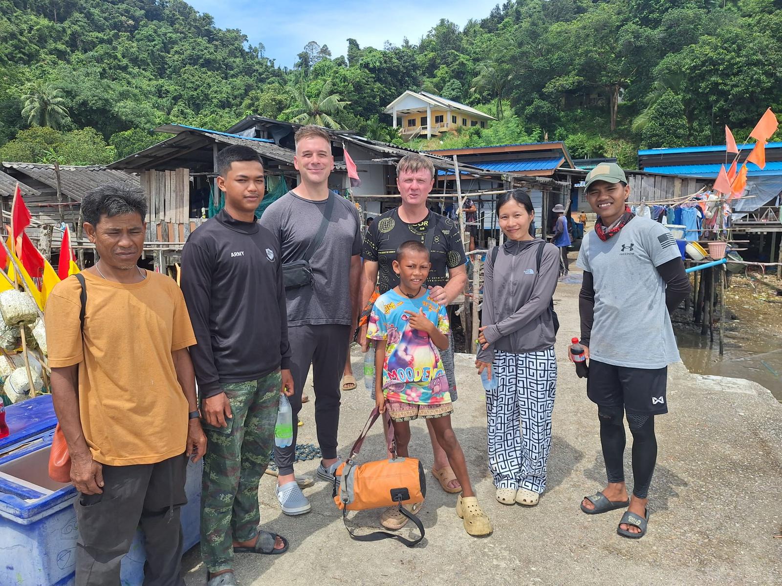 Visitors from America, 7 September: seven people, among them two Western men, local adults and a boy, on a concrete jetty in a coastal stilt-house village with flags and green hills behind