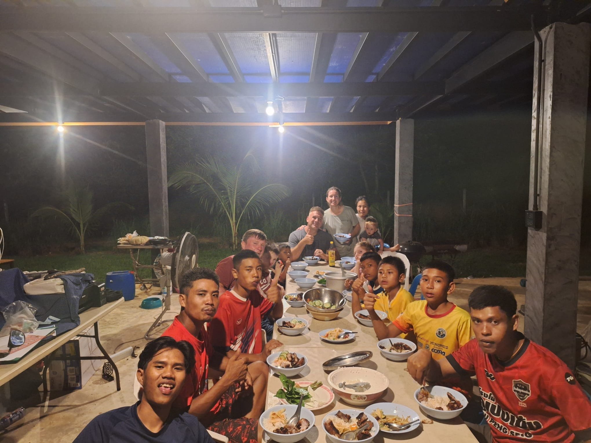 A barbecue evening with Dan and his family: teenage boys in football jerseys and visiting adults and children share a meal at a long table under a lit pavilion