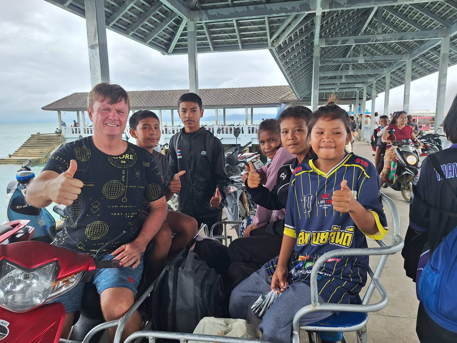 Sports week in Ranong: a Western man and several schoolchildren giving a thumbs-up from a motorbike sidecar on a pier, with the sea behind