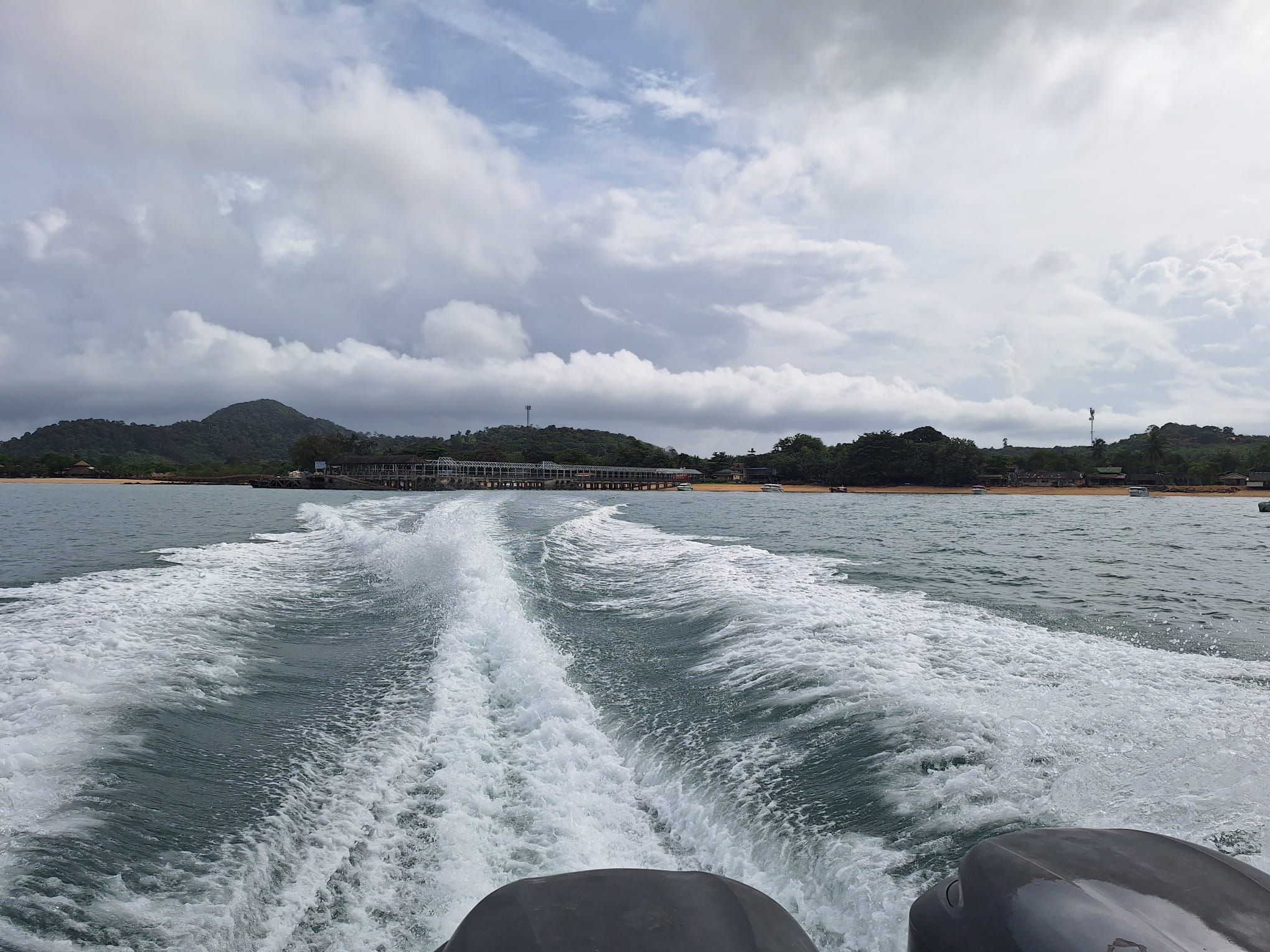 The view back from a speedboat on the way to Bangkok for the visa in August 2025: a white wake behind twin outboard motors, with the island's pier and forested hills under cloud