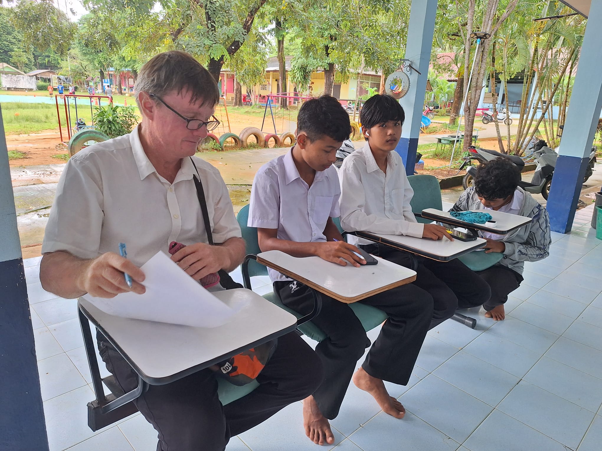 Philip writing his own exam alongside Moken students in white school shirts at tablet-arm desks on a school veranda, with the schoolyard and playground behind