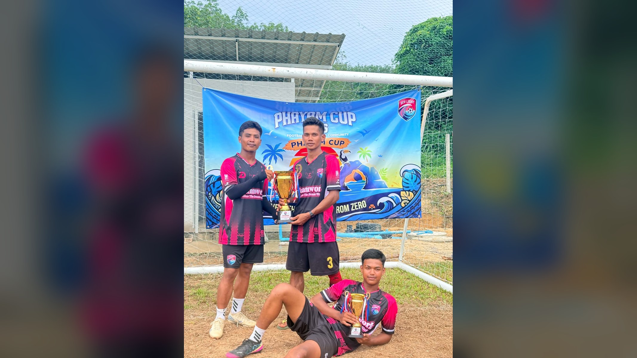Three young men in pink-and-black Sea Light 'Teamwork' kits, two standing with a large gold trophy and one lying in front with a smaller one, before a goal and the Phayam Cup banner; Sea Light B came third