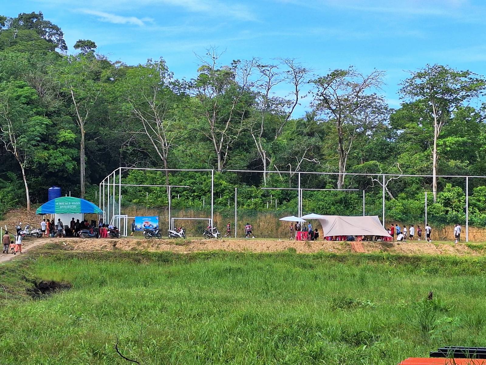 Wide view across a grassy field to the fenced soccer pitch during the Phayam Cup, with canopies, tents, parked motorbikes and spectators
