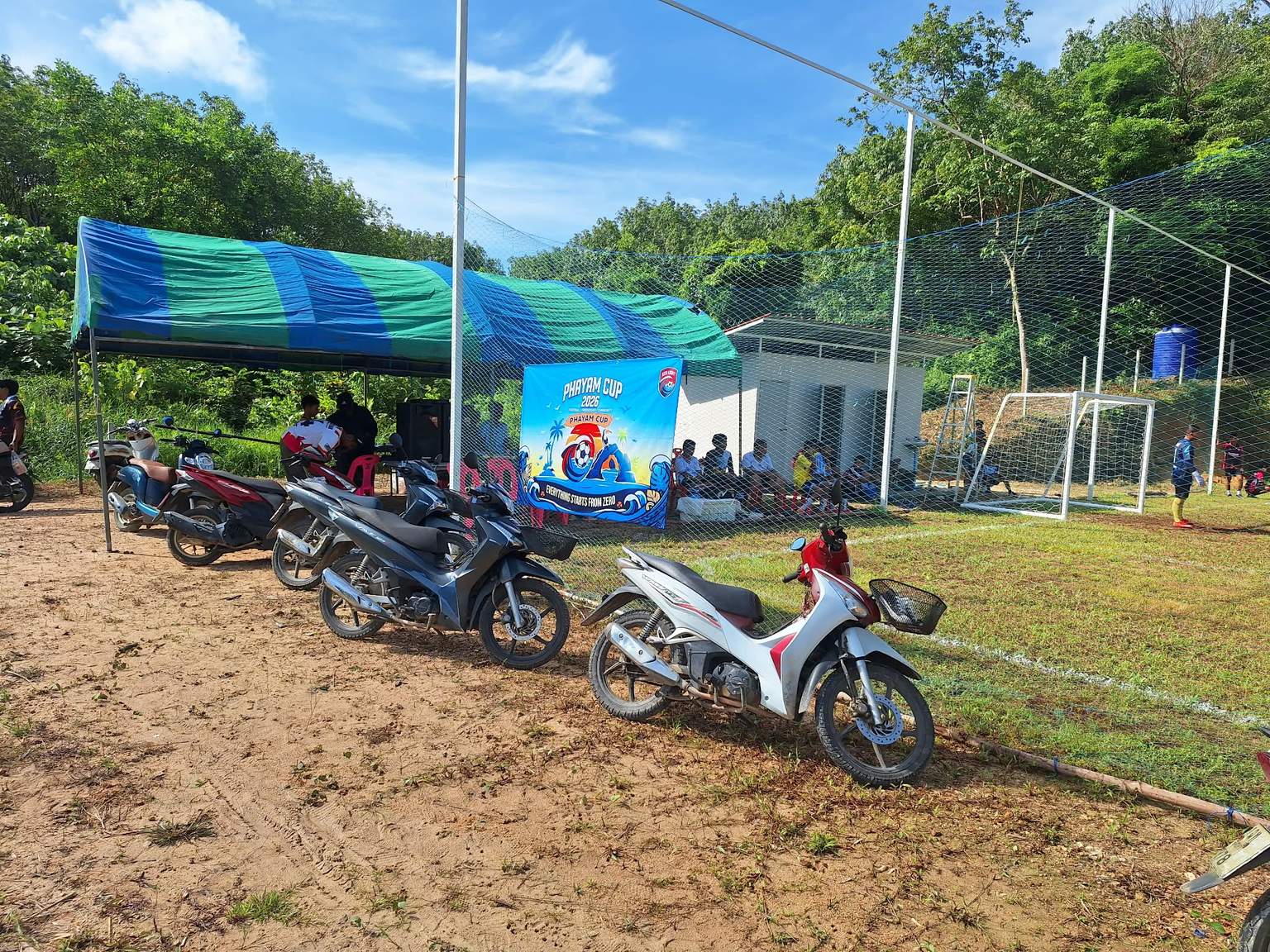 Motorbikes parked beside the netted soccer field, with a striped canopy, the Phayam Cup banner and spectators seated by a small building