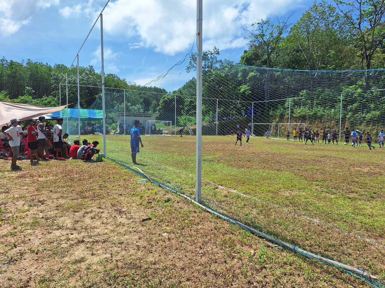 Spectators watching from under a canopy beside the netted field as players line up near the goal