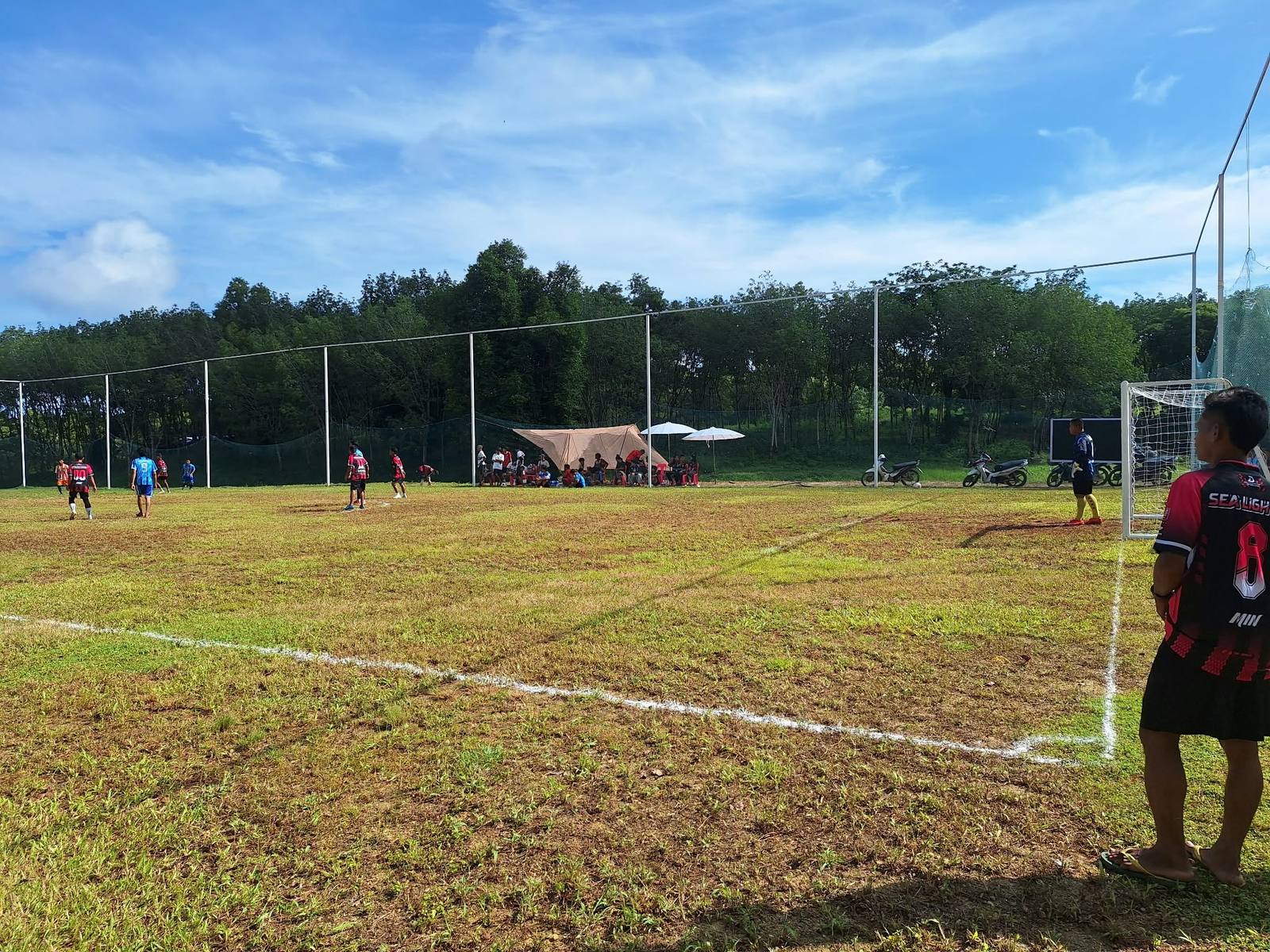 A match on the grass pitch, with a player in a Sea Light number 8 jersey watching from the touchline and spectators under a tent and umbrellas across the field