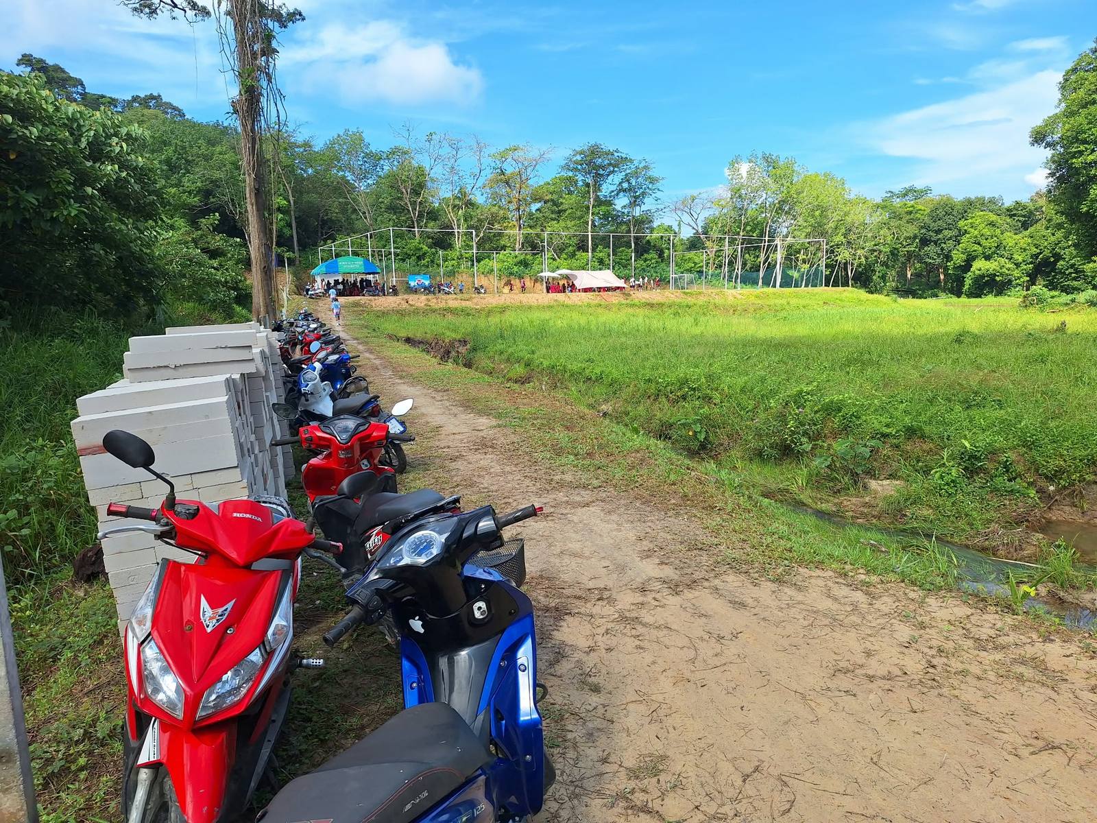 A row of motorbikes parked along the dirt track beside stacked concrete blocks, leading to the soccer field and the crowd